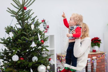 Annesi küçük oğlunu kollarında tutuyor. Çocuk parmağıyla Noel ağacının tepesini işaret ediyor. Kadın, bebeği havaya kaldırır. Tamam mı? Anne ve oğlu Noel ağacında takılıyor..