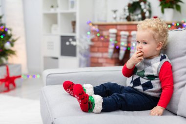 Küçük bir çocuk gri bir kanepede oturur. Çocuk ileriye bakıyor ve parmaklarını ağzına sokuyor. Arka plan bulanık. Arka planda bir Noel ağacının bir parçasını görebilirsiniz ve altında sunumlar vardır. İçeri