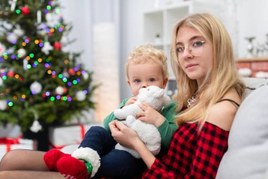 Kadın zarif bir ekose elbise giyiyor. Kadın kucağında küçük bir çocuk tutuyor. İkisi de gri bir kanepede oturuyor. Oda Noel süslemeleriyle süslenmiş. Kadın giyinmiş.