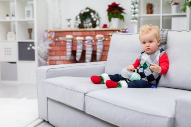 Çocuk gri bir kanepede oturuyor. Arka planda üzerinde Noel süsleri asılı bir şömine görebilirsiniz: renkli ışıklar ve Noel çorapları. Çocuk renkli bir şey giyiyor.