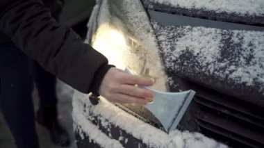 A person is shoveling snow from her car with a scraper. She scrapes the lights of her car. The lights are on. There is a lot of fresh snow lying around. It is a cloudy frosty day.
