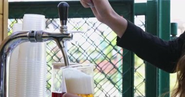- Bara yakın çekim. Barın üzerine bir musluk yerleştiriliyor. Bir kadın plastik bardağa bira dolduruyor. Çok sayıda bardak ve yanında şeker kutusu olan bir kap yanında duruyor. Konteyner ahududu içeriyor.