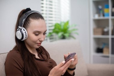 Bir kız bej bir kanepede otururken cep telefonu kullanıyor. Kızın kafasında kulaklık var. Kız kulaklıkla müzik dinliyor. Arka planda beyaz duvarlı bir pencere görebilirsiniz.