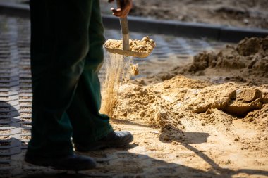 Bir inşaat işçisi elle metal bir kürekle kuru kumu taşır. Güneşli bir gün ve yüksek hava sıcaklığı dışarıda çalışırken hız kazandırmaz..
