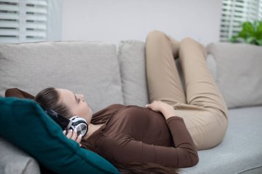 Kız bej bir kanepede uzanıyor ve bacaklarını kanepenin arkasına yaslıyor. Kızın kafasında kulaklık var. Başını turkuaz bir yastığa yaslıyor. Kız rahatlarken...