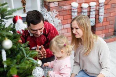 Bir kadın, bir adam ve küçük bir kız Noel ağacının yanında kalıyorlar. Küçük kız ayakta ve ailesi onun yanında çömeliyor. Kırmızı kazaklı bir adam ellerini çırpıyor. Aile gülümsüyor.
