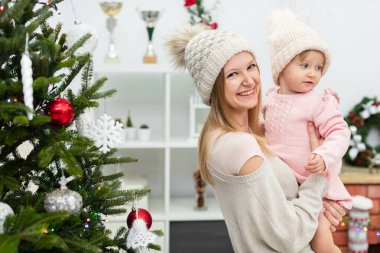 Gülümseyen bir anne ve küçük kızı tatil döneminde birlikte vakit geçirirler. Arka planda raflı beyaz bir kitaplık görülebilir. Kadının yanında süslü bir Noel ağacı duruyor.