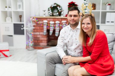 Kız ve oğlan gülümsüyor ve gri koltukta oturuyorlar. Çift, birbirini kucaklıyor. Kadın kırmızı bir elbise giyiyor ve adam desenli bir gömlek giyiyor. Arka planda görebilirsiniz.