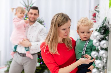 Bir anne kucağında küçük bir çocuk taşır. Kadın, çocuğa Noel çelengindeki gümüş mücevherleri gösteriyor. Arka planda bulanık bir figür görülebilir. Bir adam kucağında küçük bir kız tutuyor ve...