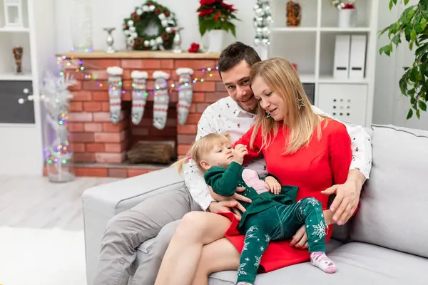 Mom and dad hug their little daughter. The whole family is sitting on a gray couch. The little girl lies on her mothers lap. The woman is wearing a festive red dress. The family is celebrating the