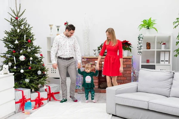 A family of three is spending the holidays in a family atmosphere. A little girl holds hands with her parents. The family stays in a room decorated with Christmas decorations