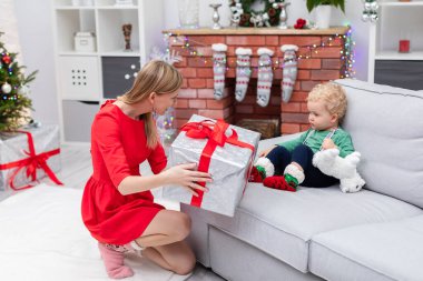 Kırmızı elbiseli bir kadın küçük bir çocuğun oturduğu koltuğun önünde çömeliyor. Anne çocuğa kırmızı kurdeleli büyük bir kutu verir. Anne çocuğuna bir Noel hediyesi veriyor.