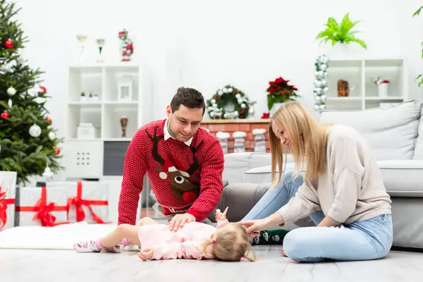 A man and a woman sit on the floor and play with their little daughter. A little girl in a pink dress lies on the floor. The room is decorated with Christmas decorations and a Christmas tree.