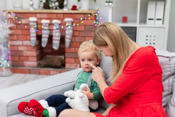 A woman in a red dress touches a little boys nose with her index finger. The child is sitting together with his mother on a gray couch. In the background you can see a fireplace decorated with