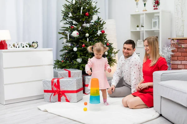 Mom and dad are sitting on the carpet, not far from the Christmas tree. Next to her parents stands a little girl. The child is wearing a pink dress. The parents are playing with the child in stacking