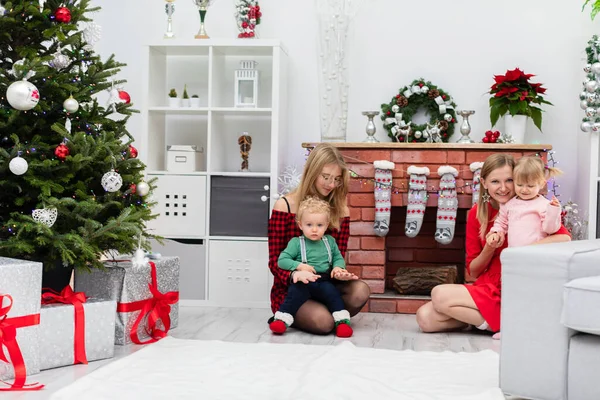 The brick fireplace is profusely decorated with Christmas decorations. Wood lies in the fireplace. A woman sits by the fireplace and hugs a little girl in a pink dress. Next to her sits another woman