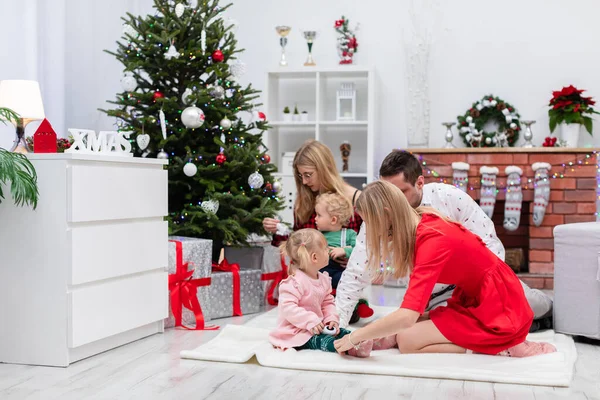 2人の女性 1人の男と2人の子供が一緒にクリスマスを過ごします 家族はクリスマスツリーでカーペットで遊んでいます 部屋はクリスマスの装飾で飾られています クリスマスツリーの下 — ストック写真