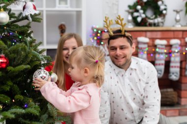 Ön planda küçük bir kız Noel ağacı mücevherlerine bakıyor. Bir çocuk elinde Noel ağacı süsleri tutar. Arka planda gülümseyen ebeveynler oturuyor. Adam gömlek ve gömlek giyiyor.