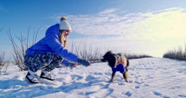 Sıkı bir ceket giymiş genç bir kız kış yürüyüşüne çıktı. Kadına renkli kapüşonlu ceket giymiş bir köpek eşlik ediyor. Sahibi köpeğiyle oynuyor ve onu okşuyor..