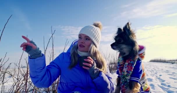 一个女人和一只狗一起坐在雪地里 天气很冷 所以这只狗穿着蒙面夹克和围巾 那只狗朝女人指指的方向看去 — 图库视频影像