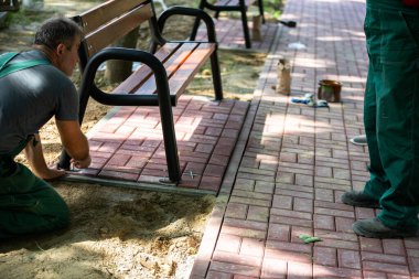 Güneşli bir günde, bir işçi parktaki bankı elle kurarken bir diğeri durup izliyor. Yaya geçidindeki yeni oturma kapları..