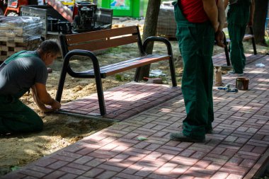 Güneşli bir günde, bir işçi parktaki bankı elle kurarken bir diğeri durup izliyor. Yaya geçidindeki yeni oturma kapları..