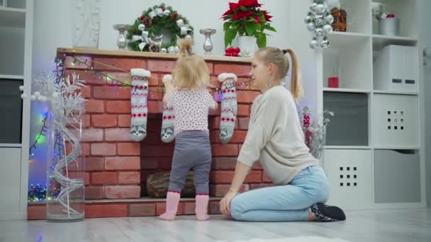 若い女性が暖炉でひざまずき 小さな娘と遊んでいる 女の子は暖炉のそばに立って クリスマスの装飾を見ています ある時点で 子供は吊るすライトをつかみ — ストック動画