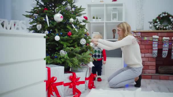 一个年轻的女人跪在圣诞树旁 把装饰品挂在树枝上 一个好奇的小男孩走在圣诞树旁 可以看到一个用彩灯装饰的砖壁炉 — 图库视频影像