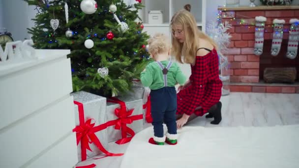 クリスマスツリーでエレガントな模様のドレスを着た若い女性 彼女の隣には小さな男の子が立っている 女性はクリスマスツリーの下に横たわっているリボンと結ばれた子供の大きな箱を示しています — ストック動画