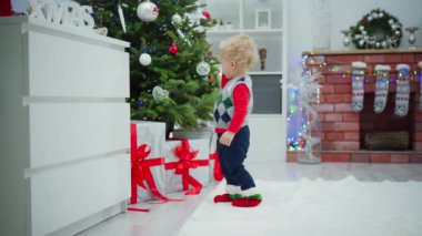 Süslü bir yelek giymiş küçük bir çocuk süslü bir Noel ağacına doğru yürüyor. Kırmızı kurdelelere sarılı büyük hediye kutularının orada bir an için duruyor..