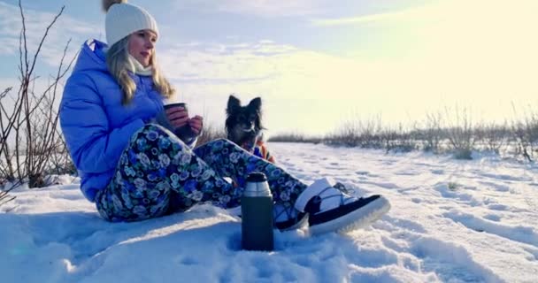 雪の中に座っている少女が熱い飲み物を飲んでいます 明るい冬の日で あちこちに雪がたくさんあります 遠くに荒野が広がっている 女の子の隣に座っている小さな — ストック動画