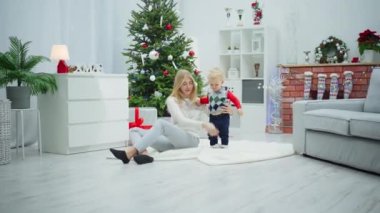 Uzun saçlı sarışın bir kadın ve beyaz halıda oturan küçük bir çocuk. Halı odanın ortasında, Noel süslemeleriyle dolu. Bir noktada, kadın yere uzanır.