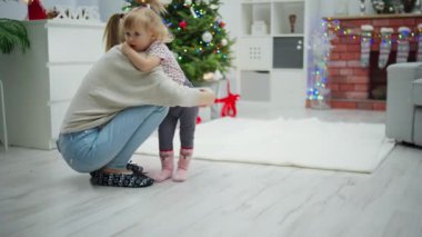 Genç bir kız küçük bir kızın yanına çömelmiş ve onu kucaklıyor. Kadın çocuğa sıkıca sarılıyor ve şefkatle yanağından öpüyor. Büyük, süslü bir Noel ağacı ve diğer tatil süslemeleri...