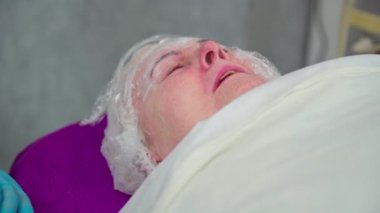 Yaşlı bir kadın yatakta iki kez hapşırır. Kadın bir güzellik salonunun müşterisi. Yüzü kısmen folyoya sarılı, saçı da örtülü. Kadın kozmetik tedavi görüyor..