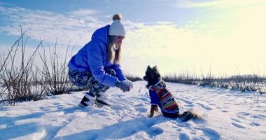 Kız karda çömeldi ve elinde bir avuç dolusu kar topladı. Karla oynuyor, etrafa saçıyor. Kızın önünde renkli bir ceket giymiş küçük bir köpek oturuyor. Köpek bakıyor.