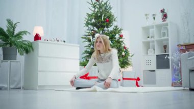 Gözlüklü genç sarışın bir kadın odanın ortasında beyaz bir halıda oturuyor. Odada birçok Noel süsü ve büyük bir Noel ağacı var. Kadın bacak bacak üstüne atmış bir şekilde oturur.