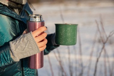 Bir kadının elinin yakın plan çekimi. Bir elinde termal bardak, diğer elinde normal bir kupa tutuyor. Kadın açık bir ceket ve açık parmaklı eldivenler giyiyor..