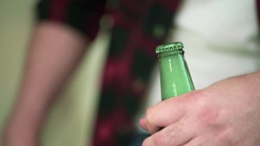 Yeşil bir şişenin boynundaki yakın çekim. Bir adam cam bir şişe tutuyor. Şişeyi bir açacağıyla açıyor. Beyaz köpük ortaya çıkar..
