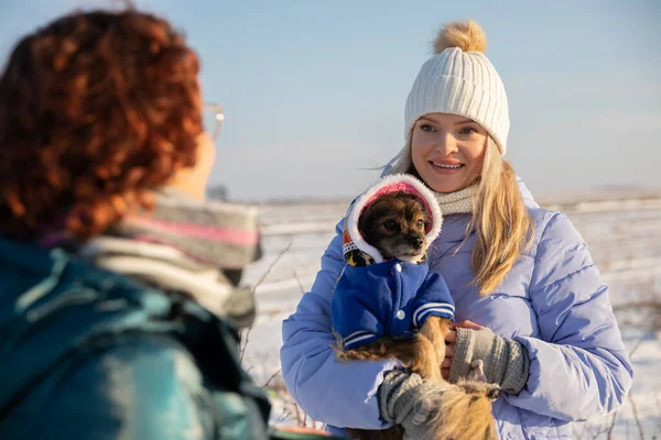 Blurred foreground. In the foreground you can see the figure of a woman standing with her back. In the background stands a smiling blonde woman holding a small dog in her arms. Snow-covered fields can
