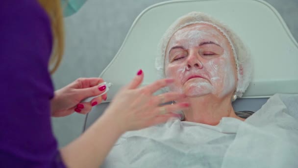 美容师的特写手将产品应用到病人脸上 美容师用润肤霜滋润一位老年妇女的脸部 这个女人头上戴着一顶保护帽 — 图库视频影像