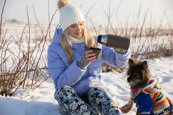 Expedition with a dog in the wilderness. The winter weather makes it necessary to warm up. A girl sits in the snow and pours herself tea from a thermos. Next to her sits a small dog.