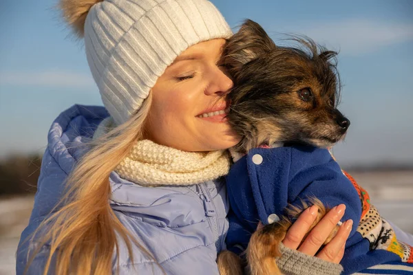 Close-up of the face of a smiling young woman. She cuddles her face into the fur of the dog she is holding in her arms. The woman and dog are wearing warm jackets.