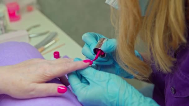 两名妇女的手的特写 美容师正在用深色粉红的光油给她的客户刷指甲油 在背景中 你可以看到一个台面上站着化妆品器皿 — 图库视频影像