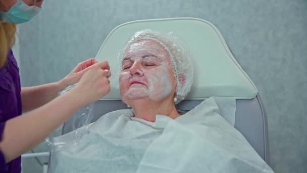 一个女人正坐在一张化妆品椅上 她把眼睛闭上了 她的脸上涂了一层厚厚的白霜 美容师把胶卷贴在女人的脸上 — 图库视频影像