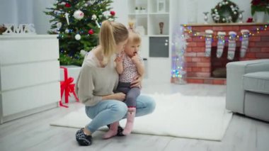 Küçük bir kız annesinin dizine oturuyor. İkisi de birbirlerine sarılıyorlar. Bir noktada beyaz halıya şakayla sarılıp uzanıyorlar. Arka planda bir Noel ağacı görebilirsiniz.