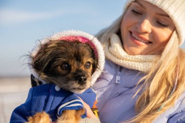 Genç sarışın bir kadın küçük köpeğine bakarken gülümsüyor. O, köpeği kollarında tutuyor. Kadın ve köpek dışarıdalar, dondurucu soğuk varken..