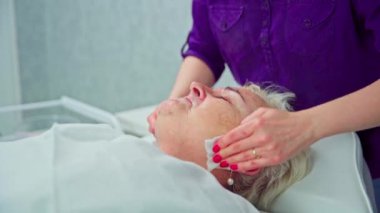 Güzellik uzmanı, müşterisinin yüzünü estetik ameliyatlara hazırlamak için derisini dikkatlice siler. Bir kadın güzellik salonundaki bir yatakta gözleri kapalı yatıyor..