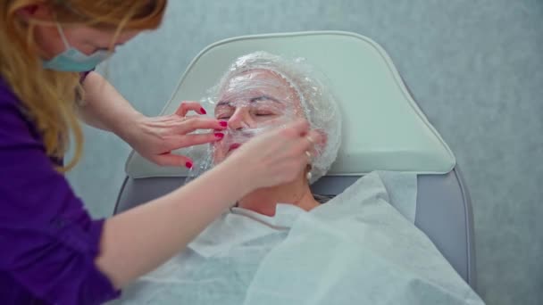 化粧品学者は 患者が皮膚に適用される準備がより効果的に機能するように特別なフィルムで顔を包みます 患者はヘッドレストと椅子に座り 目を閉じています — ストック動画