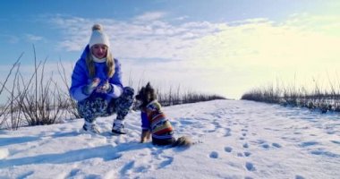 Karda bir kış yürüyüşü. Bir kız kardan ellerini sıkar. Önünde bir köpek oturuyor. Köpek kapüşonlu renkli bir ceket giyiyor..