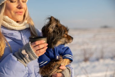 Sahibi tarafından kollarında tutulan küçük bir köpeğin yakın çekimi. Kadın diğer elinde sıcak bir içecek olan bardağı tutarken köpeğe sarılıyor..